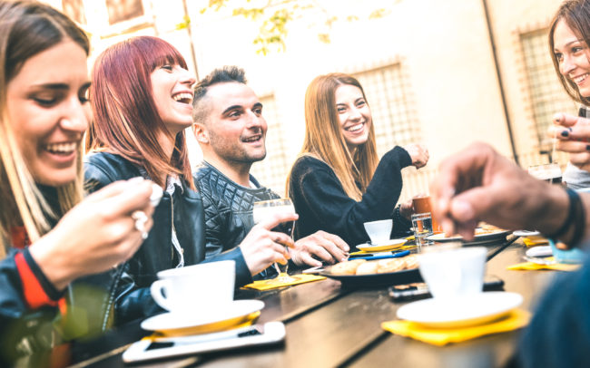 Gruppo di amici seduti a un tavolo all’aperto che ridono e bevono cappuccino
