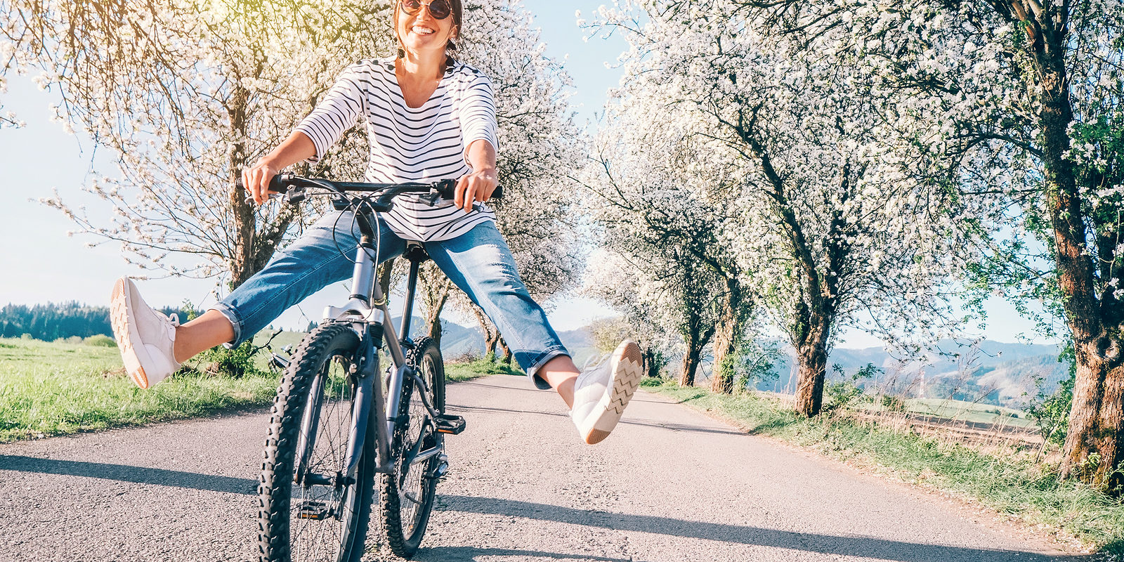 Donna felice in sella a una bicicletta su una strada circondata da alberi in fiore, con le gambe sollevate per formare un gesto di entusiasmo