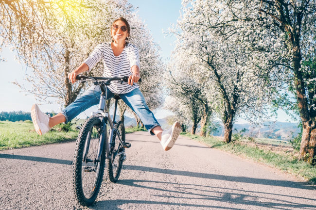 Donna felice in sella a una bicicletta su una strada circondata da alberi in fiore, con le gambe sollevate per formare un gesto di entusiasmo