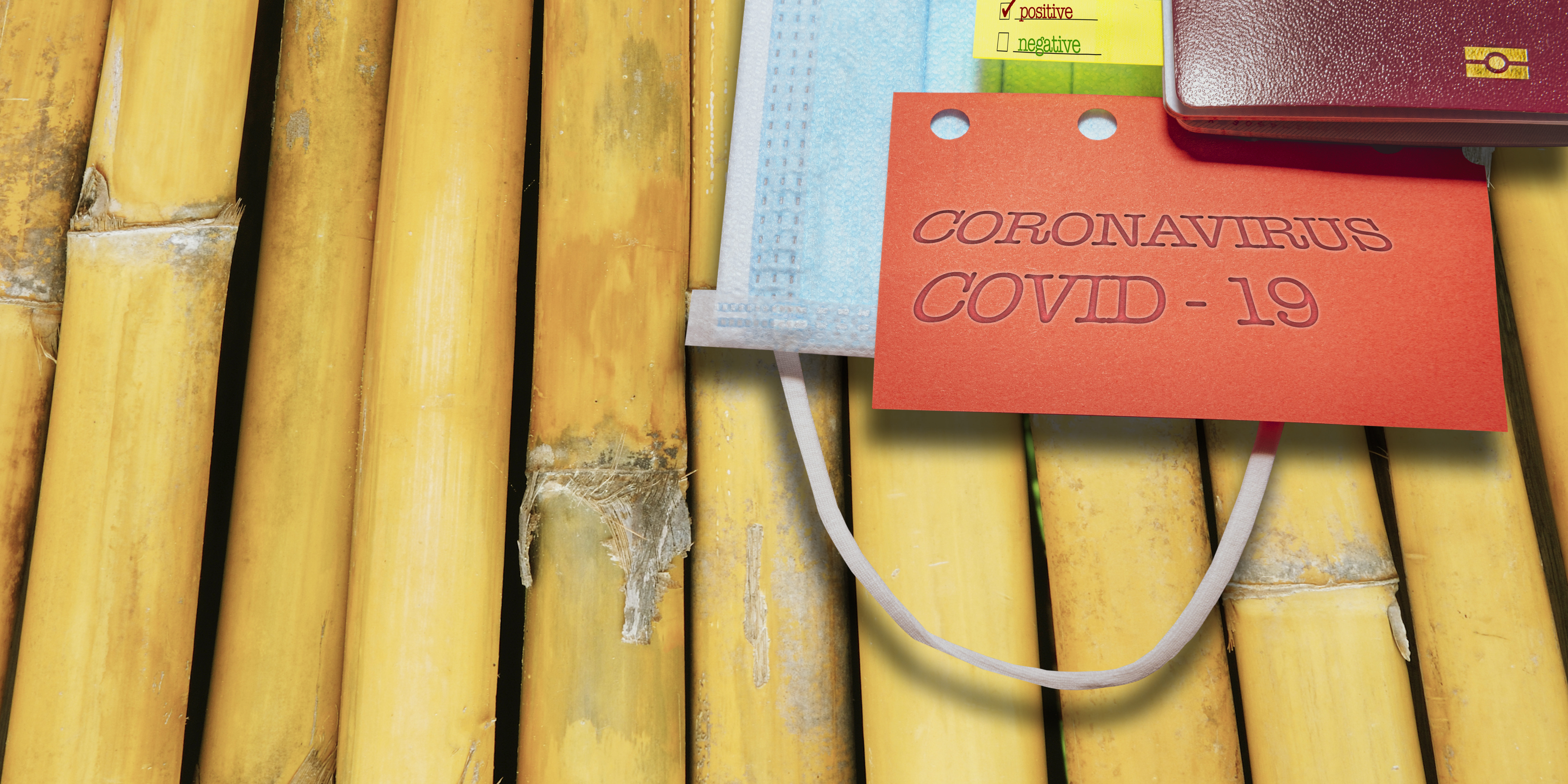 Passaporto, mascherina e cartellino COVID-19 Passaporto e mascherina chirurgica su sfondo di canne di bambù, con cartellino rosso riportante la scritta ‘Coronavirus COVID-19’ e un post-it con la casella ‘positive’ spuntata